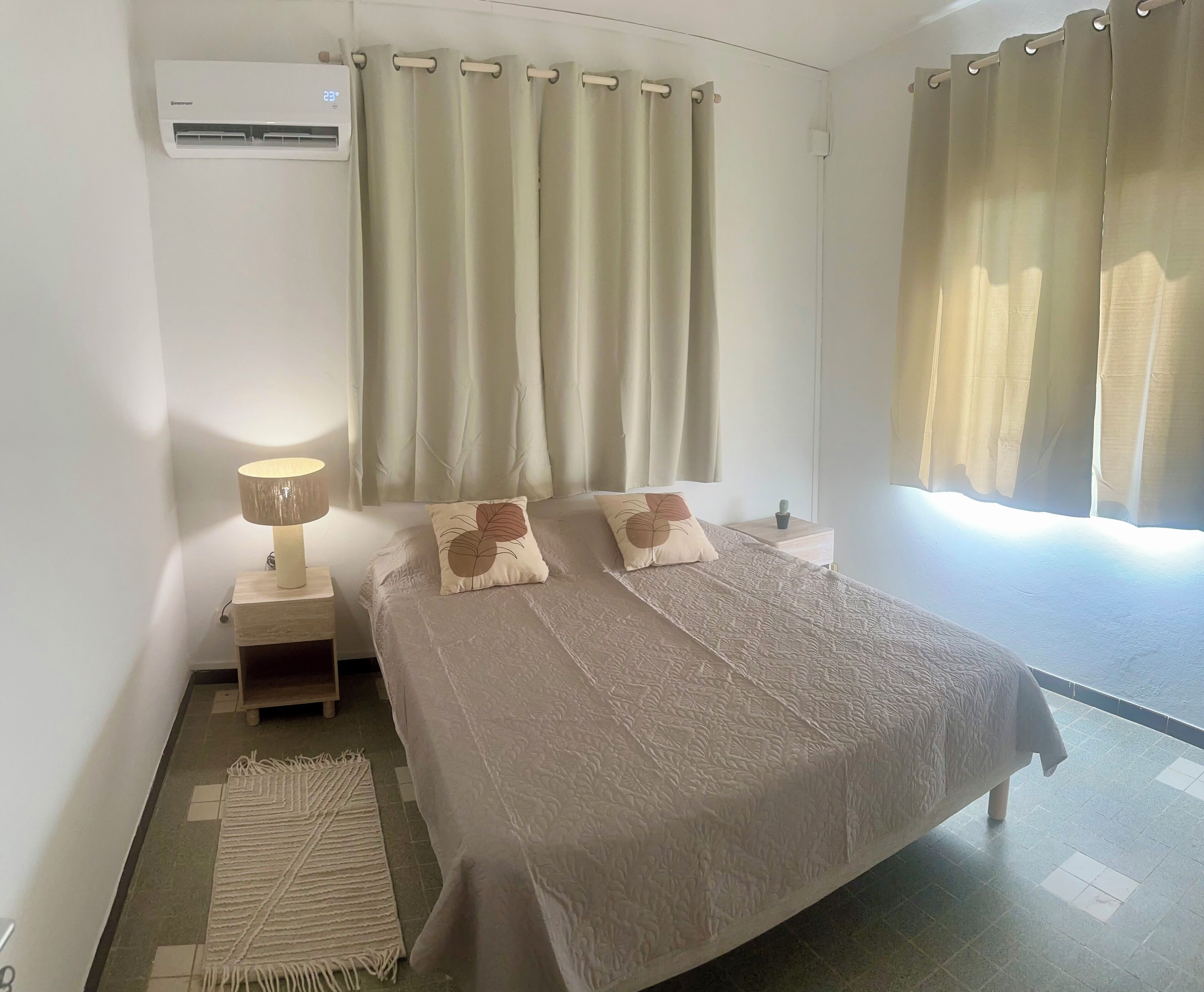 Un dormitorio con cama, lámpara de noche, mesita de noche, una pequeña planta en maceta y ventana con cortinas.