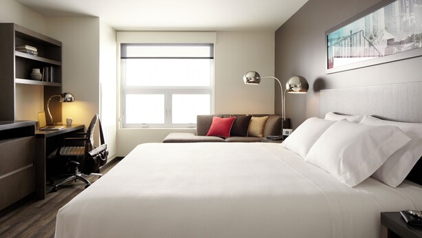 Ein Hotelzimmer mit einem großen Bett, einem Schreibtisch mit Stuhl, einem Bücherregal und einem Fenster, das für natürliches Licht sorgt.