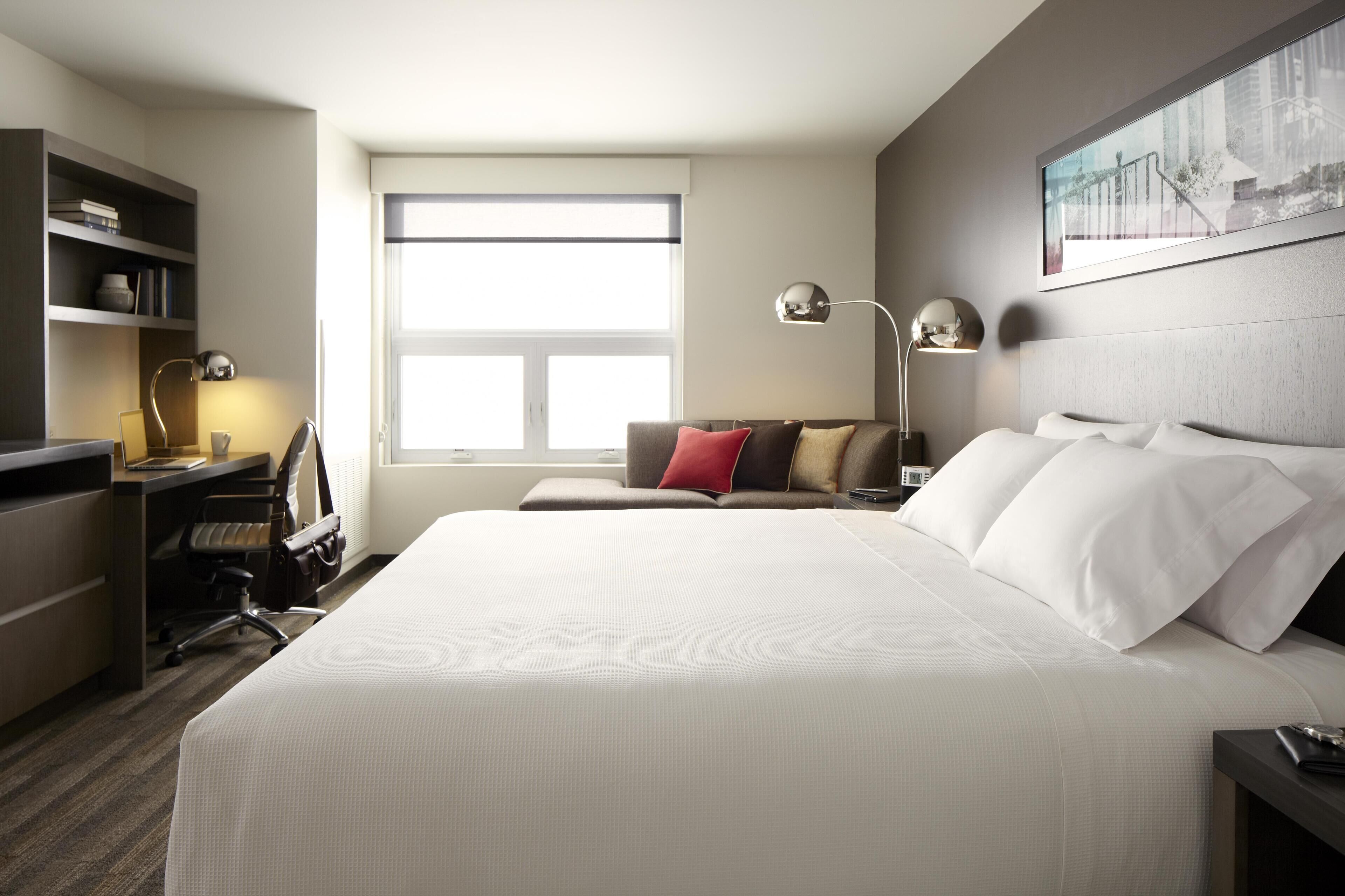 Habitación de hotel con una cama grande, un escritorio con silla, una estantería y una ventana con persianas.