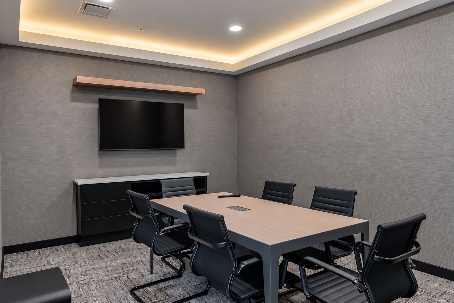 A conference room with a long table, six chairs, a flat-screen TV mounted on the wall, and a built-in desk with drawers.
