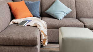Ein Wohnzimmer mit einem grauen Sofa, zwei Kissen, einer Decke und einem Buch. Über der Couch hängt ein gerahmtes Bild.