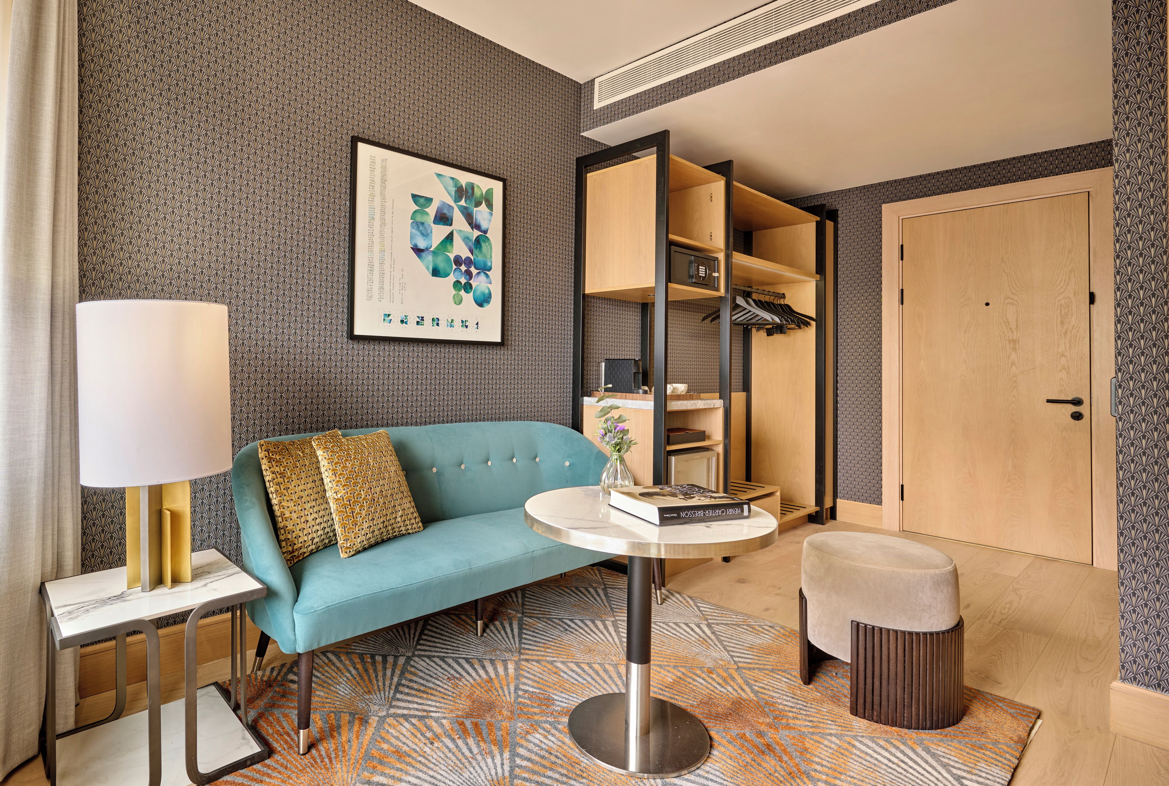 A modern hotel room with a teal sofa, a round table, a lamp, and a wardrobe.