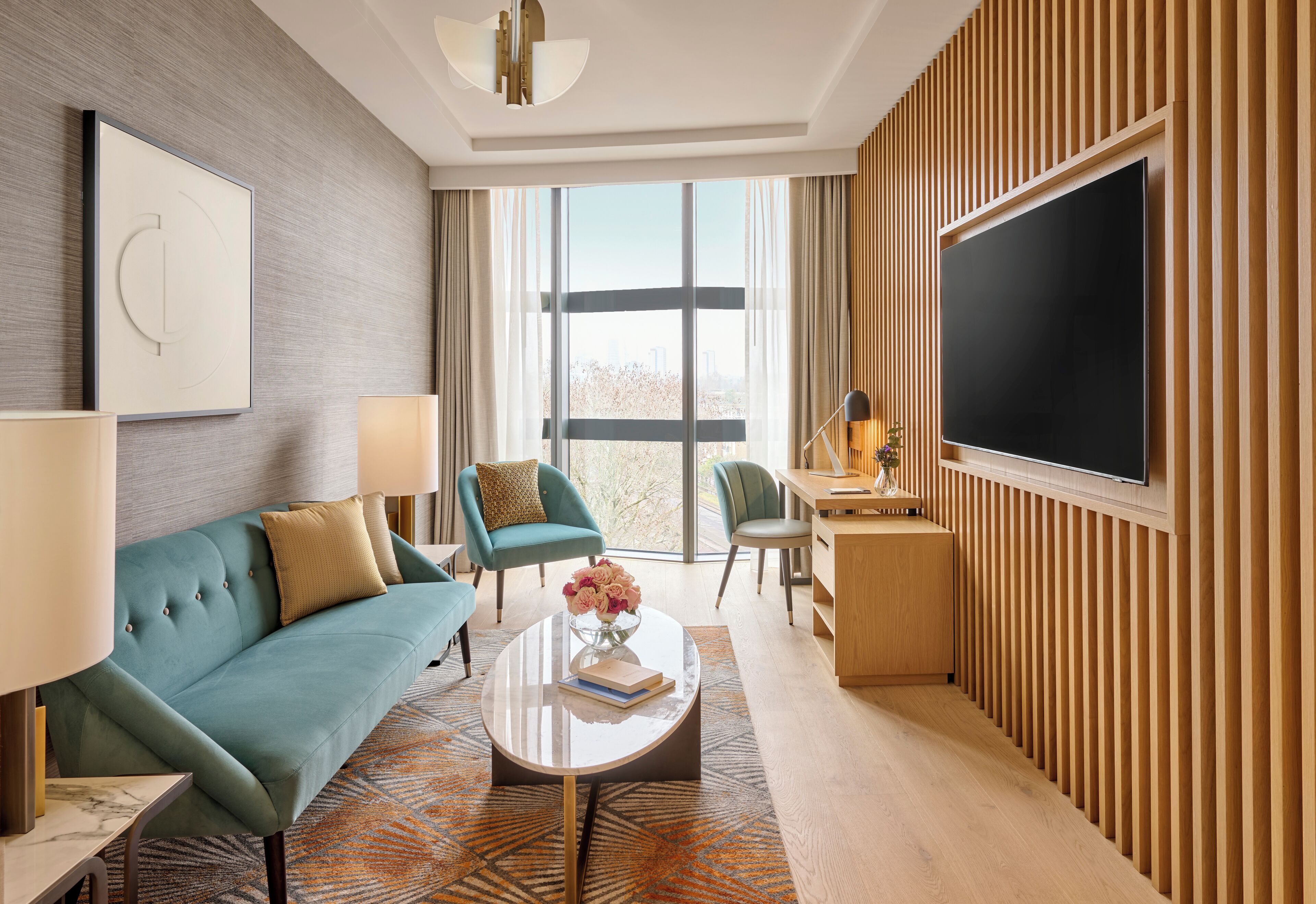 Ein modernes Hotelzimmer mit einem petrolfarbenen Sofa, einem Couchtisch mit Buch und Blumen, einem Fernseher und einem großen Fenster mit Blick auf die Stadt.