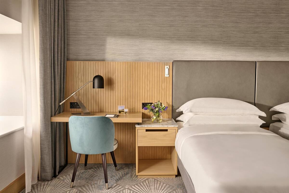 A hotel room with a bed, a desk with a chair, a lamp, and a nightstand with a vase of flowers.