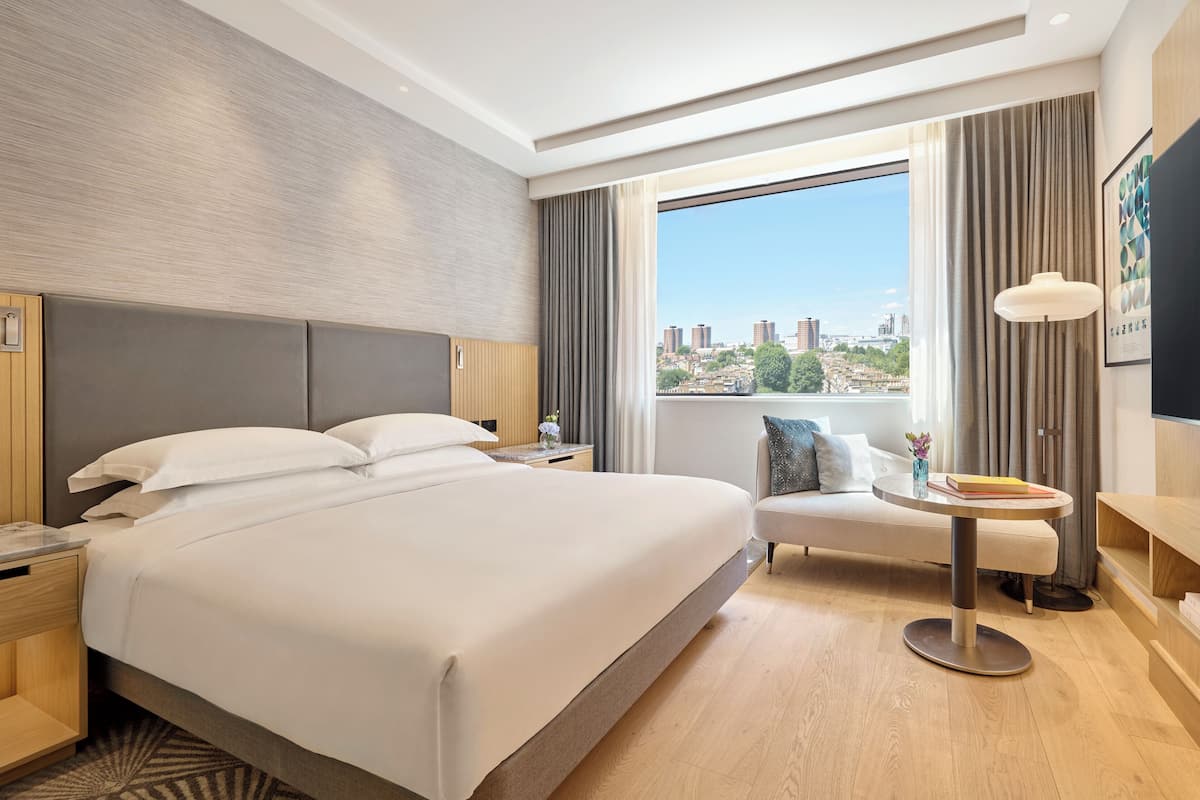 A hotel room with a large bed, a small round table, a sofa, a TV, and a window with a city view.