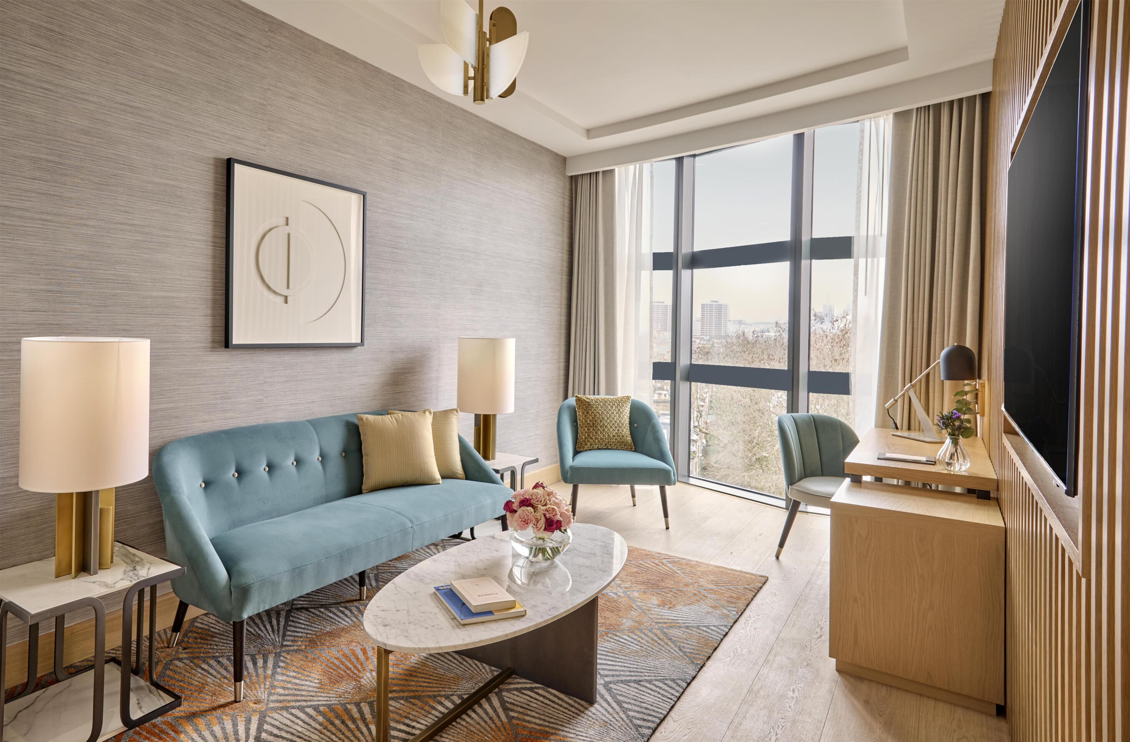A modern hotel room with a blue sofa, two armchairs, a coffee table, and a large window offering a city view.