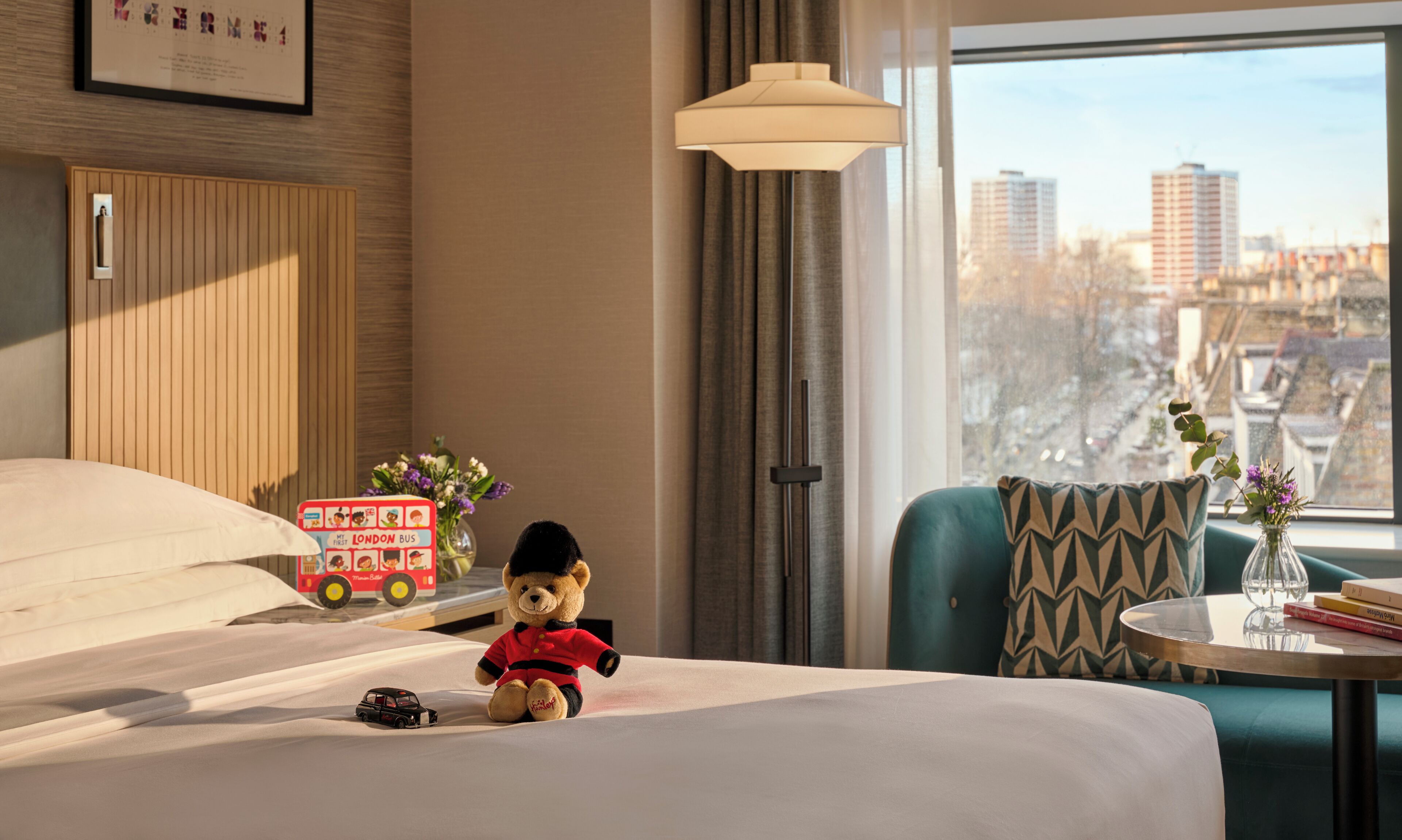 A hotel room with a bed, a teddy bear, a toy car, a vase with flowers, and a view of the city through the window.