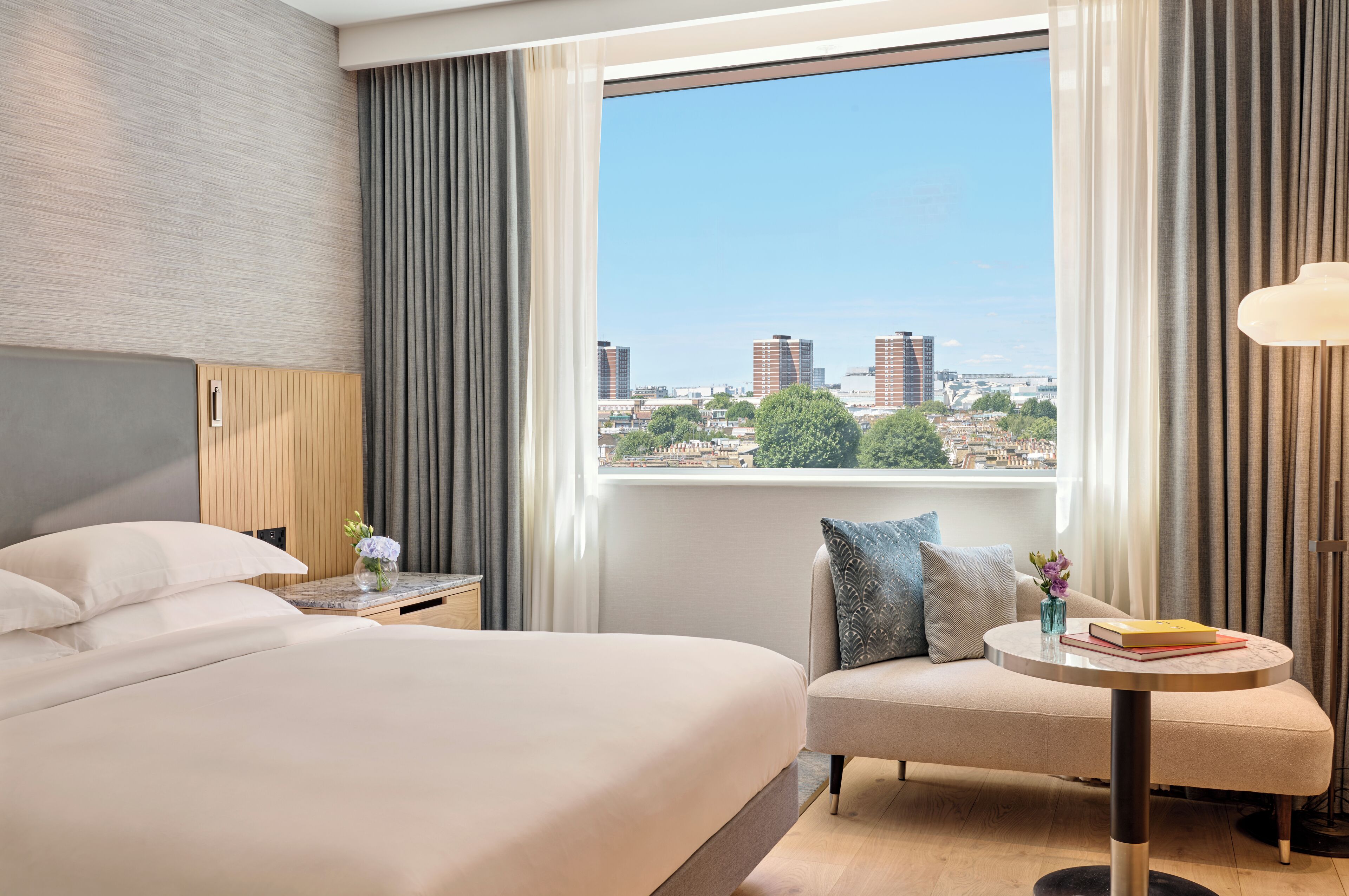 Habitación de hotel con cama, sofá, una mesita y vistas a la ciudad a través de amplios ventanales.