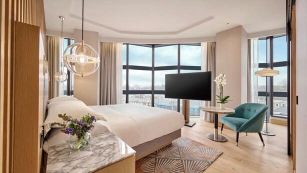A modern hotel room with a large bed, a TV, a small table, a chair, and a view of the city through large windows. - Hyatt Regency London Olympia (London)