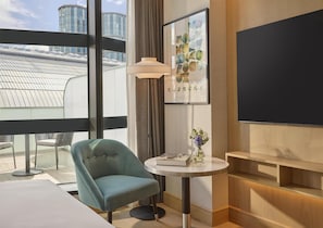 A modern hotel room with a large window, a teal armchair, a small round table with a vase of flowers, a floor lamp, and a flat-screen TV. - Hyatt Regency London Olympia (London)