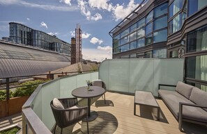A rooftop terrace with a glass balustrade, a round table with chairs, a sofa, and a view of modern buildings in the background. - Hyatt Regency London Olympia (London)
