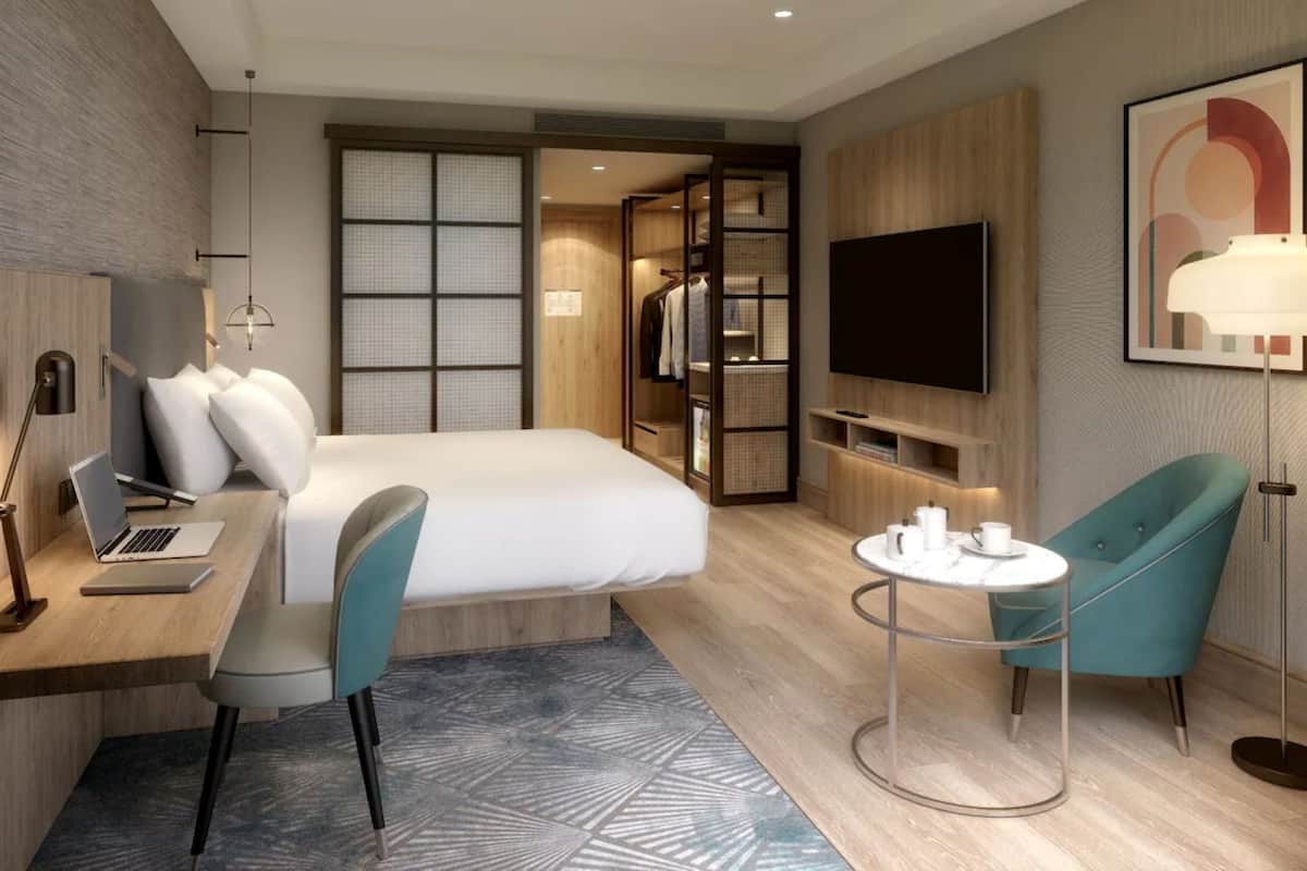 A modern hotel room with a bed, desk, chair, TV, and a small round table with a lamp.