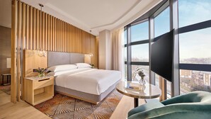 A modern hotel room with a large bed, a wooden slat wall, a round table, and a view of the city through large windows. - Hyatt Regency London Olympia (London)