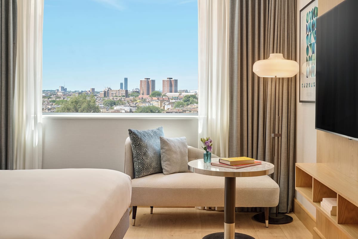 A hotel room with a bed, a sofa, a small table with books, a vase with flowers, and a view of a city skyline through large windows.
