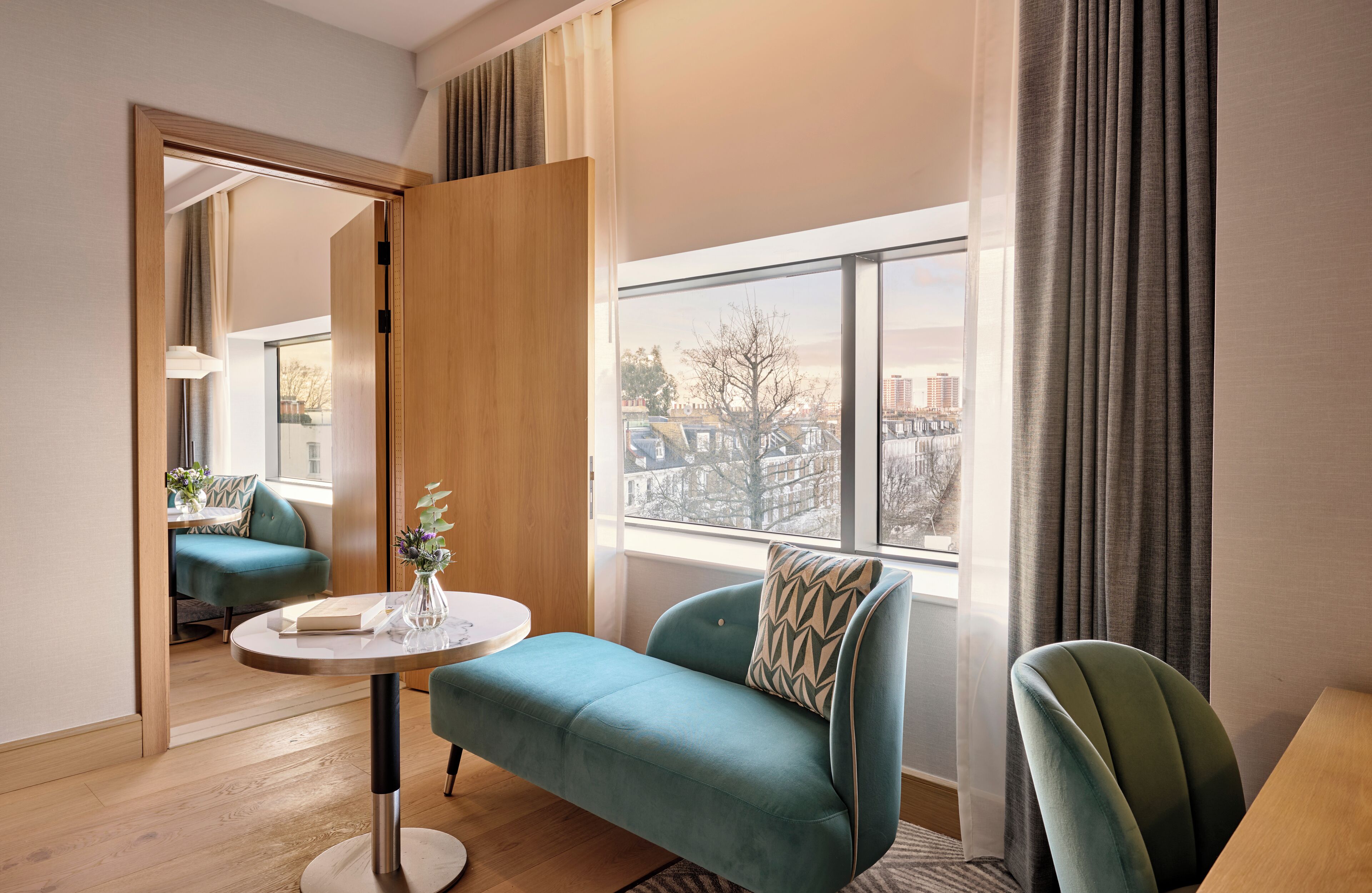 A modern hotel room with a teal sofa, a round table, and a view of the city through large windows.
