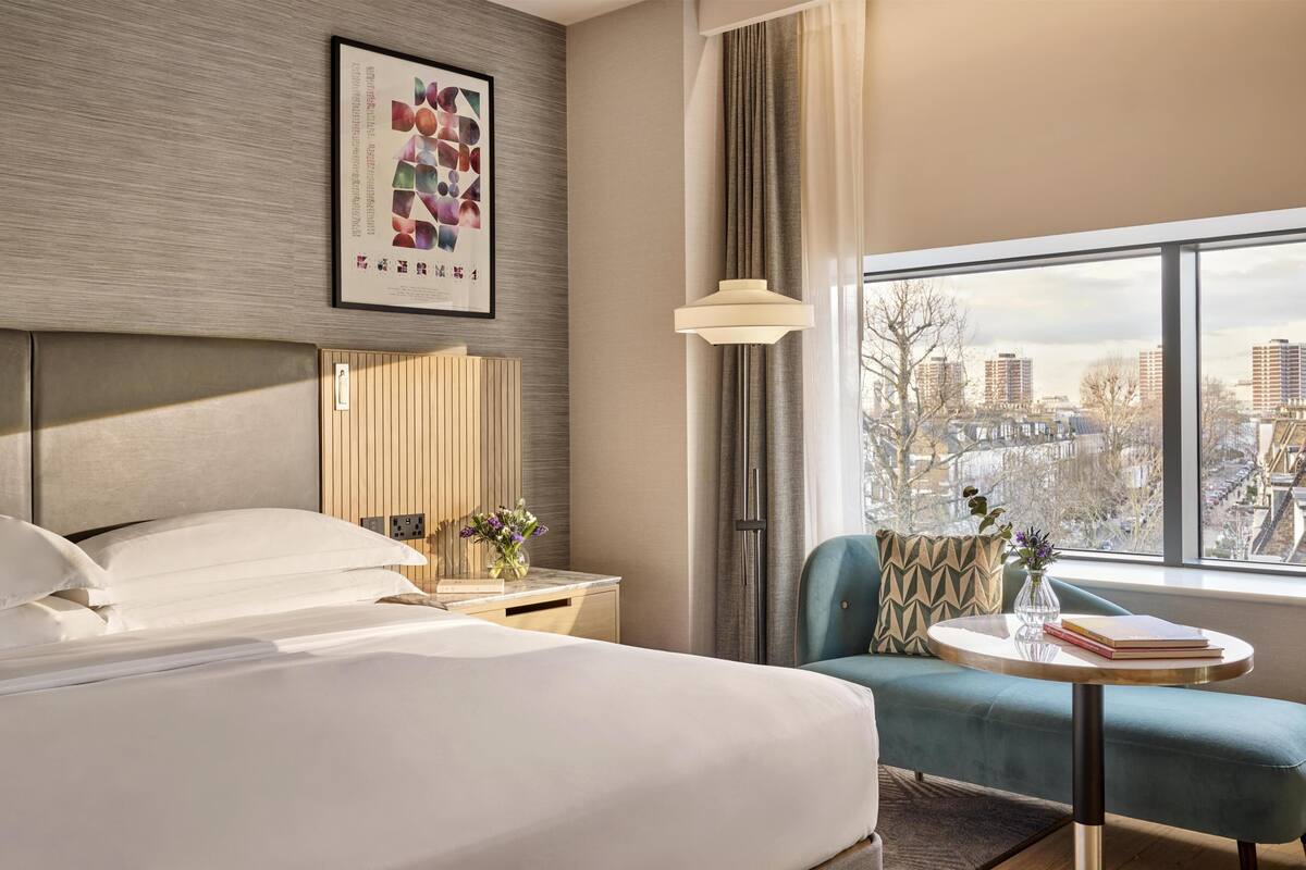 A hotel room with a bed, a seating area with a table, a city view, and a framed artwork on the wall.