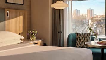 A hotel room with a bed, a nightstand with a vase of flowers, a table with books, a chair, and a view of the city through large windows.
