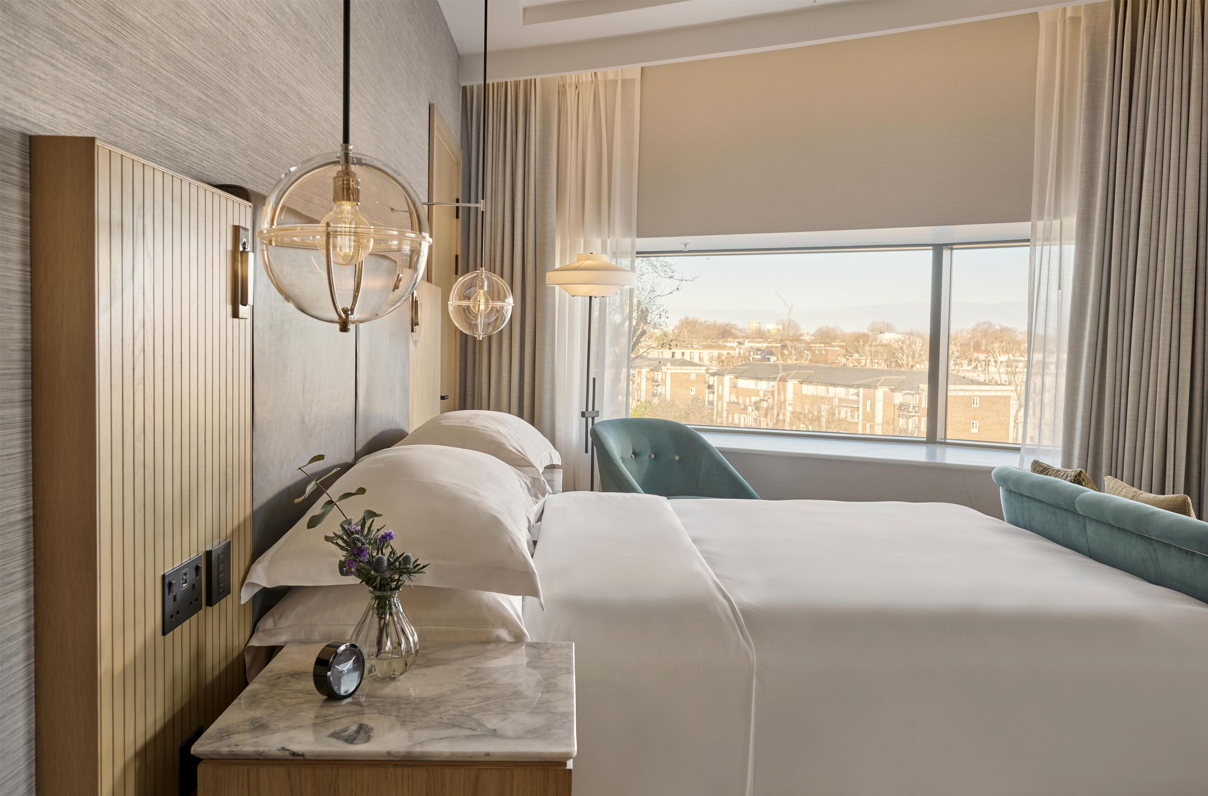 A hotel room with a large bed, a desk with a vase of flowers, a clock, and a view of the city through the window.