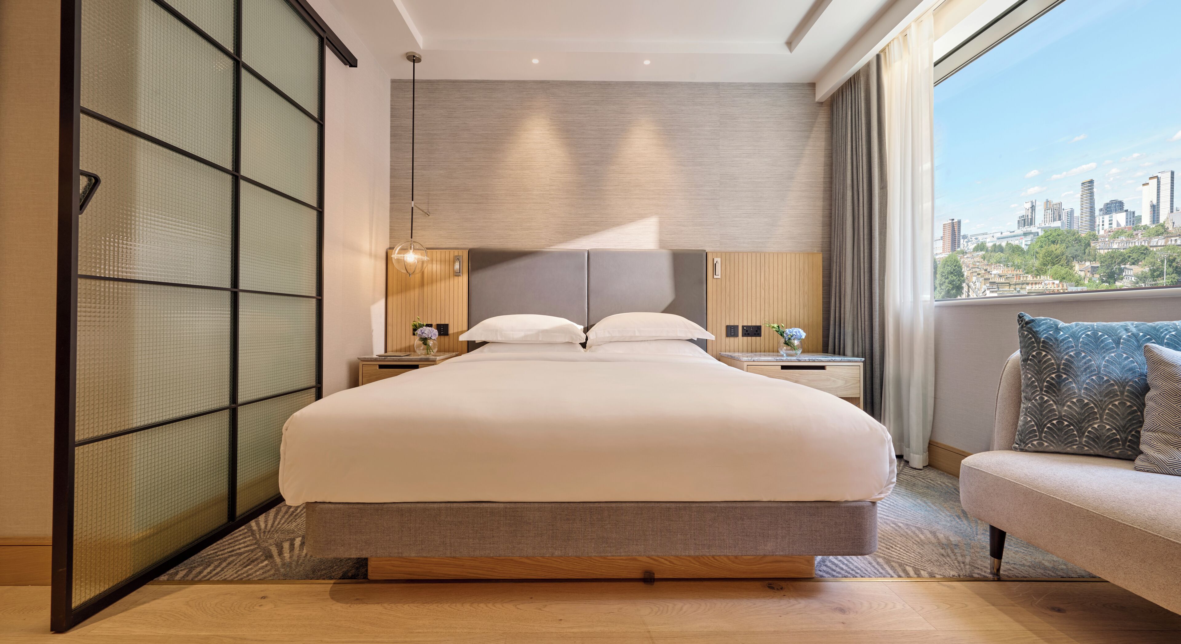 A modern hotel room with a large bed, a nightstand with a lamp, a sofa with decorative pillows, a view of the cityscape, and a sliding shoji screen.