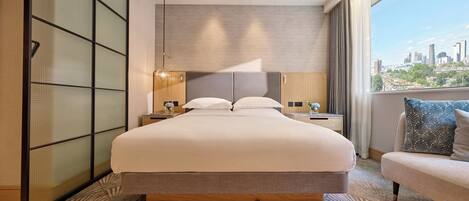 A modern hotel room with a large bed, a nightstand with a lamp, a sofa with decorative pillows, a view of the cityscape, and a sliding shoji screen.