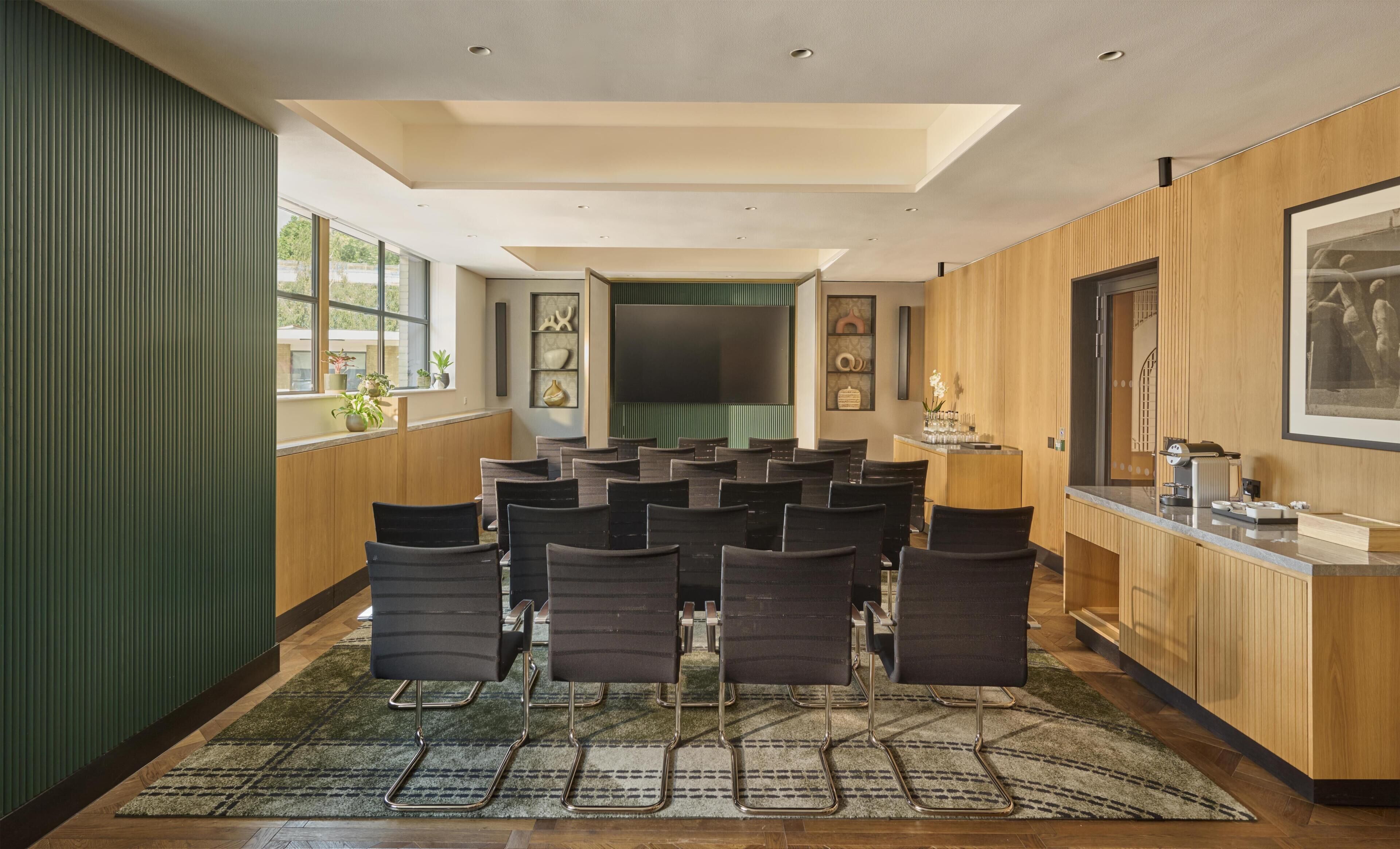 Uma sala de conferências com tela grande, diversas cadeiras, painel de parede de madeira e área de recepção com balcão.