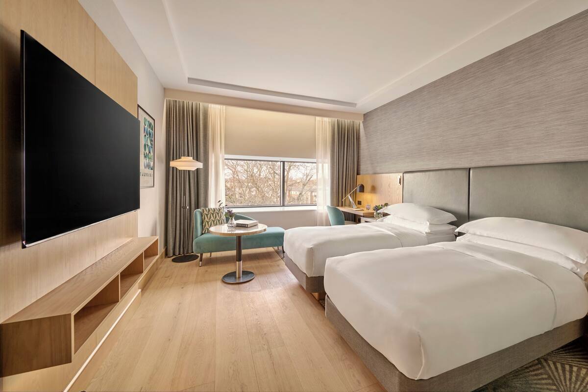 A hotel room with two beds, a TV, a desk, a chair, and a window with curtains.