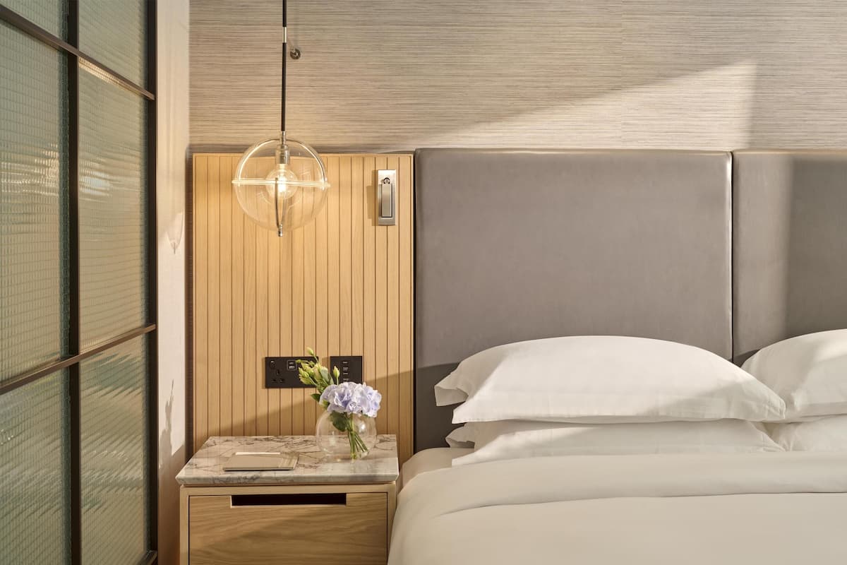 A modern hotel room with a wooden headboard, a marble nightstand, a vase with flowers, and a bed with white linens.