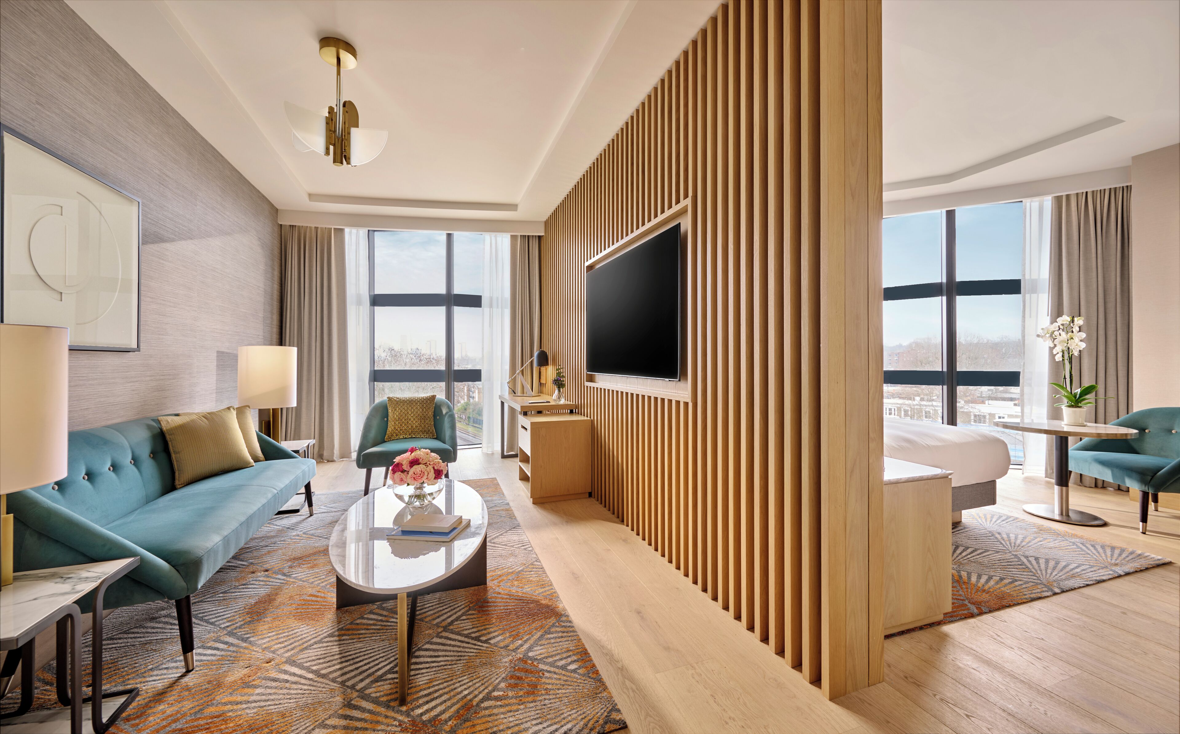 Une chambre d’hôtel moderne dotée d’une grande télévision, de canapés turquoise, d’une table basse et d’une grande fenêtre donnant sur la ville.