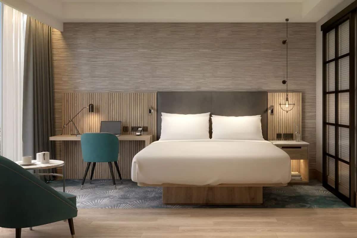 A modern hotel room with a large bed, a desk with a chair, a small table with a chair, and a large window.