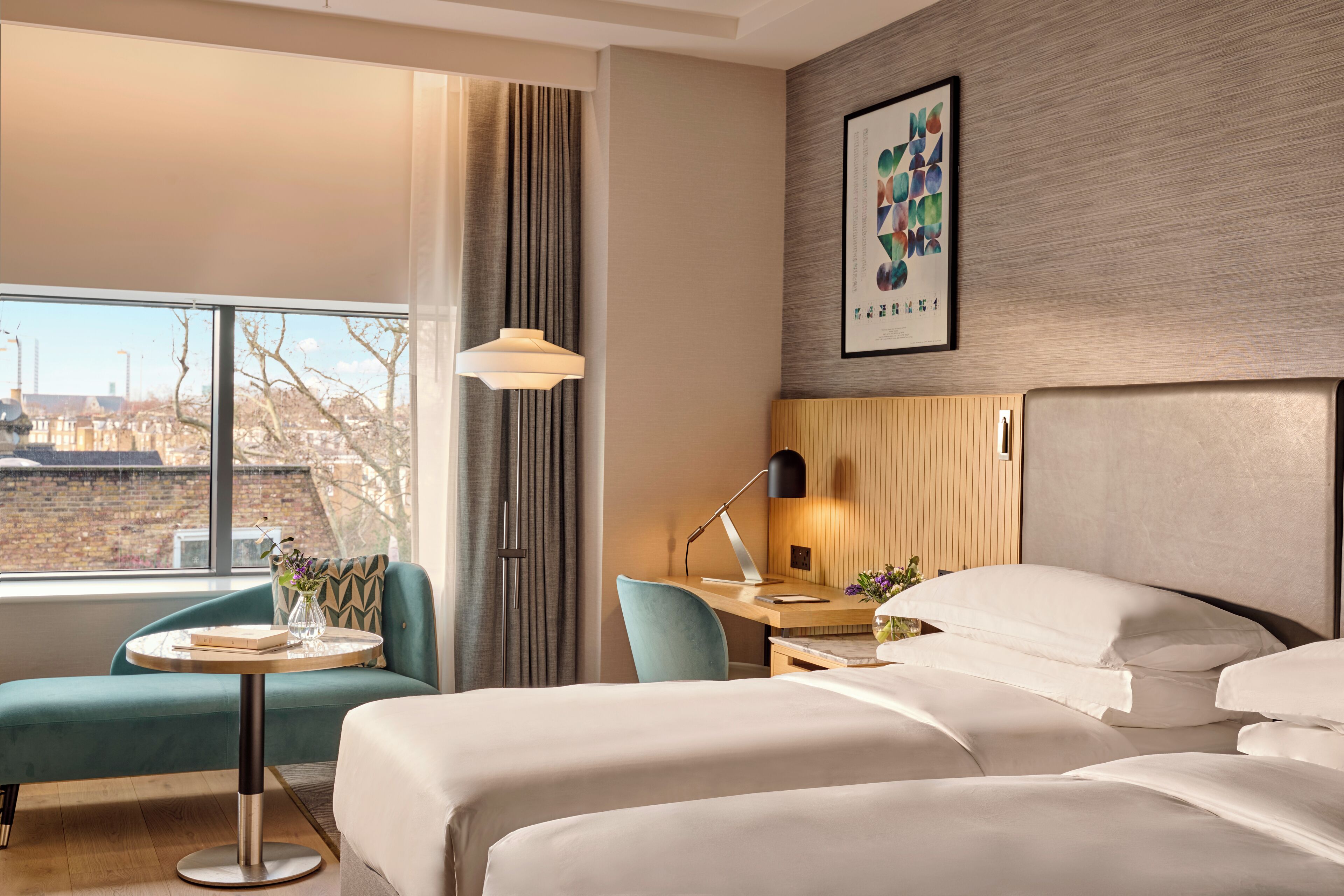 Habitación de hotel con una cama grande, un escritorio con lámpara, una pequeña mesa redonda con un florero y vistas a la ciudad a través de amplios ventanales.