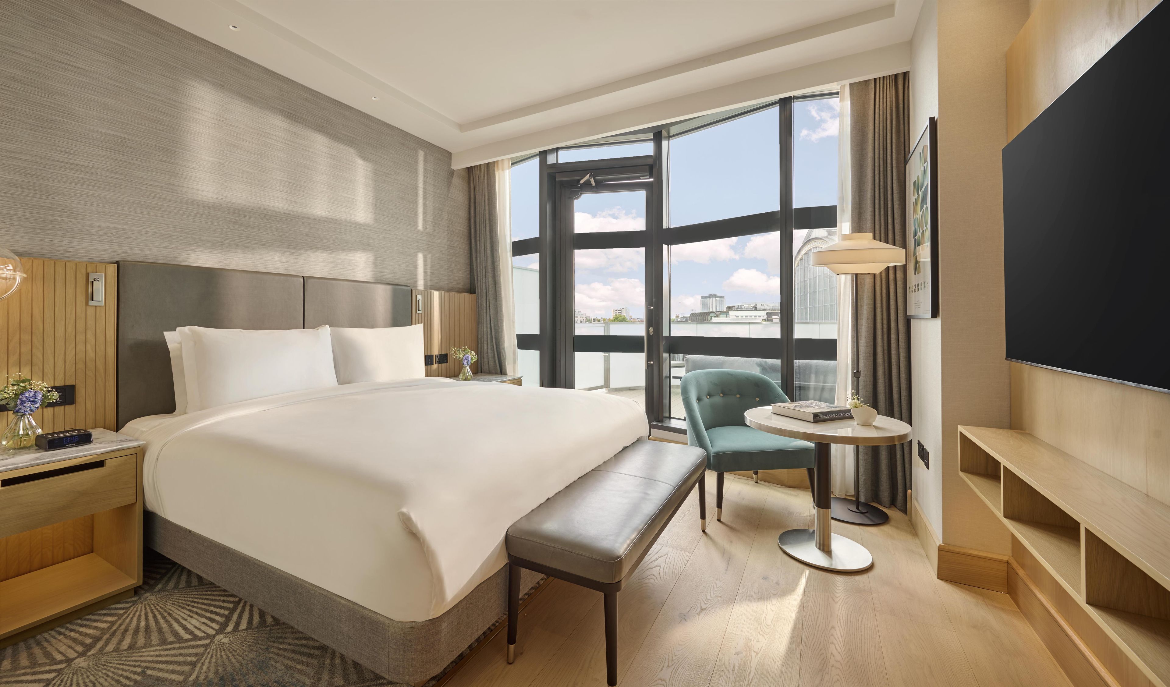 A hotel room with a large bed, a bench, a small table, a TV, and a view of the city through large windows.