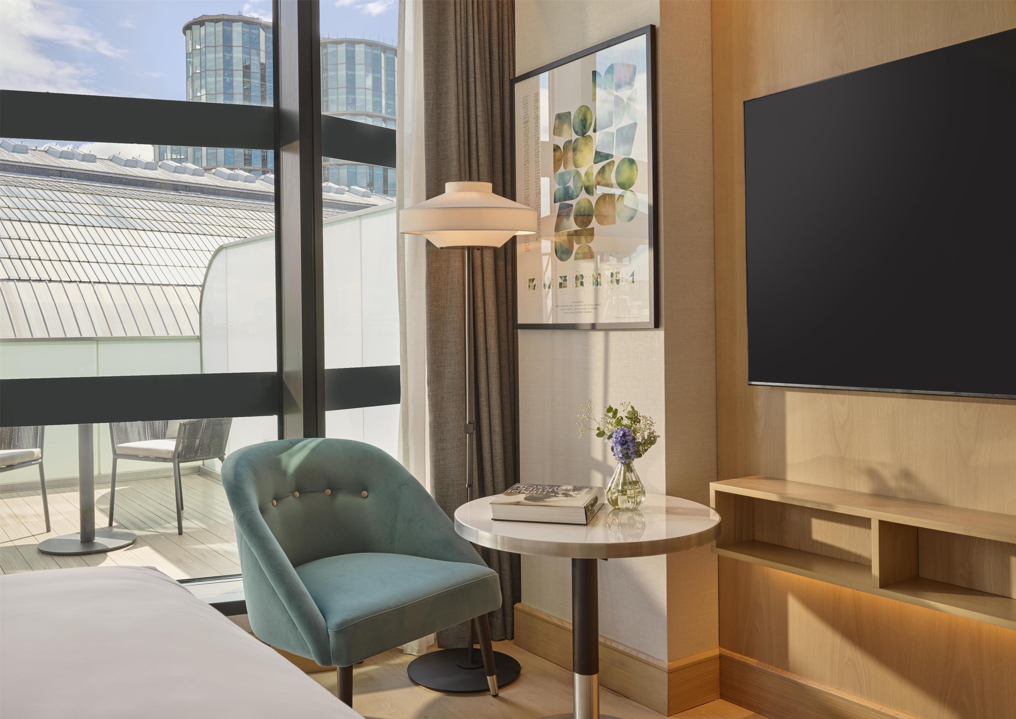 Quarto de hotel moderno com uma janela grande, uma poltrona azul-esverdeada, uma pequena mesa redonda com um vaso de flores, um abajur e uma televisão de tela plana.