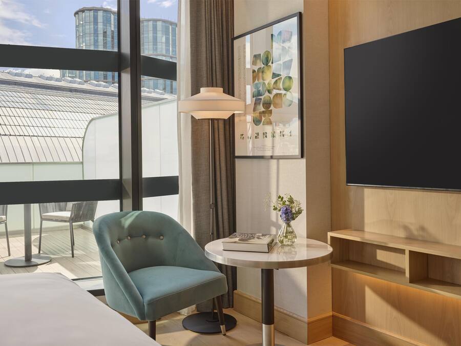 Quarto de hotel moderno com uma janela grande, uma poltrona azul-esverdeada, uma pequena mesa redonda com um vaso de flores, um abajur e uma televisão de tela plana.