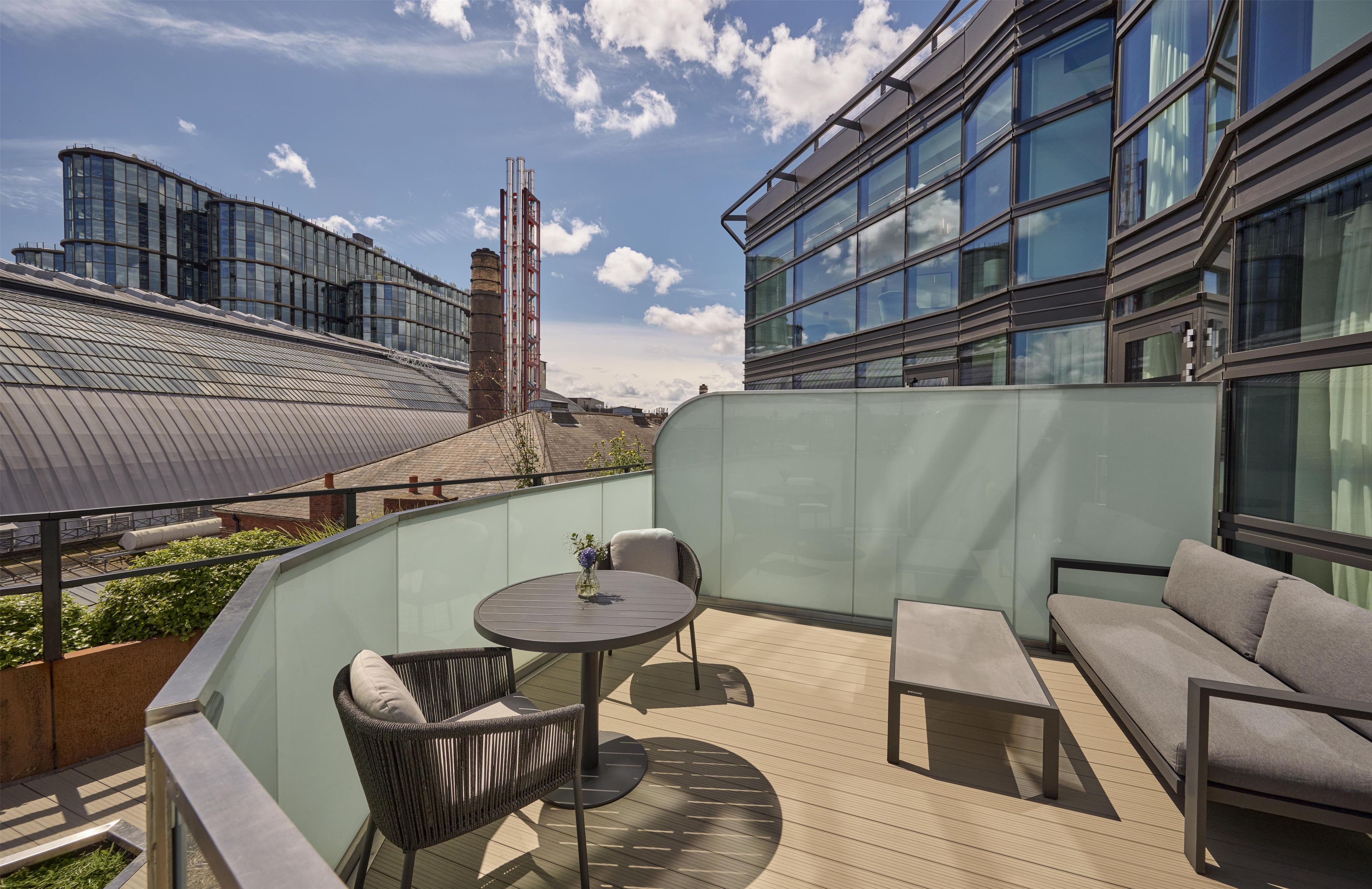 A rooftop terrace with a glass balustrade, a round table with chairs, a sofa, and a view of modern buildings in the background.