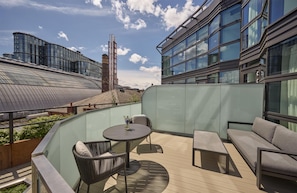 A rooftop terrace with a glass balustrade, a round table with chairs, a sofa, and a view of modern buildings in the background. - Hyatt Regency London Olympia (London)