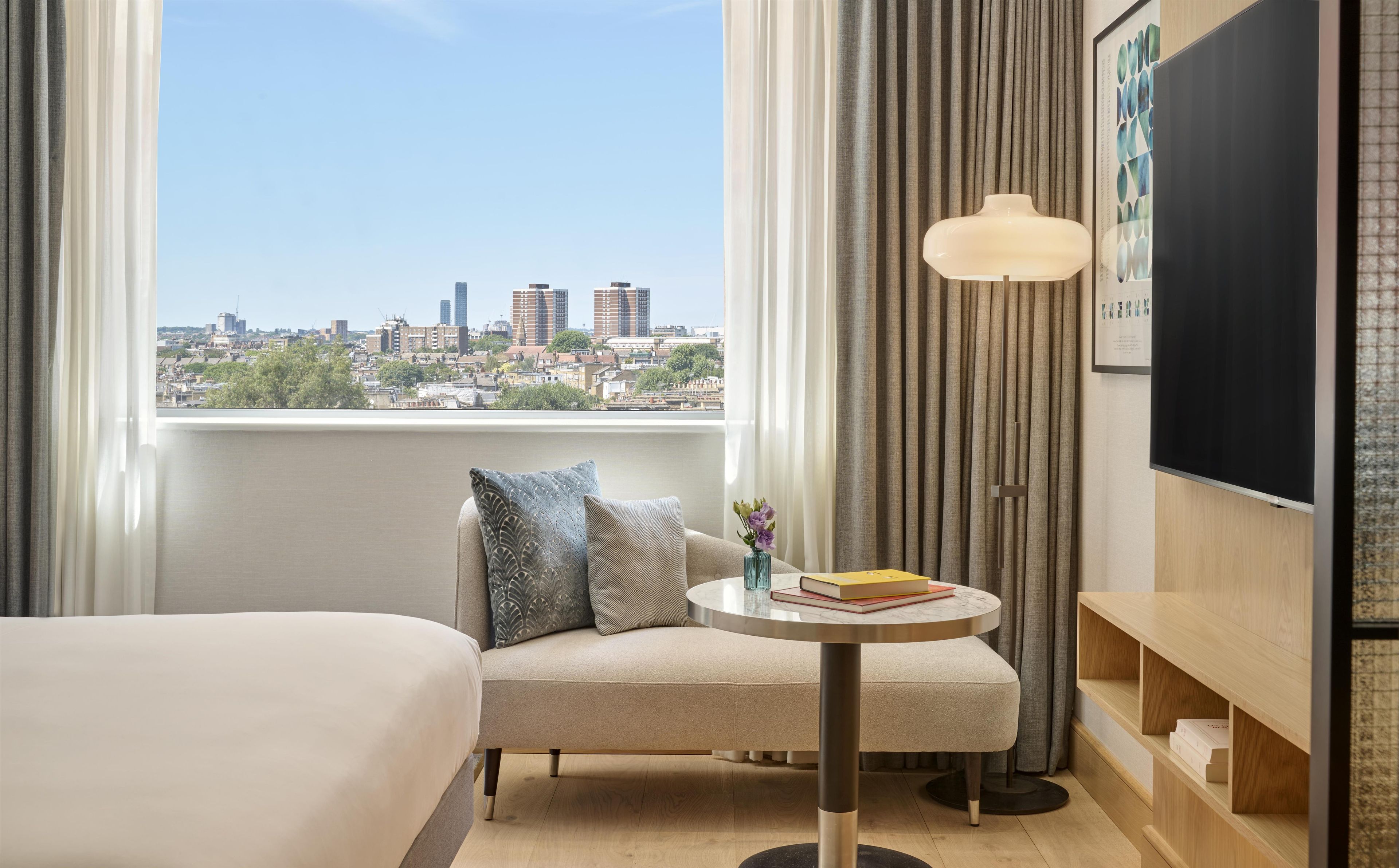Ein Hotelzimmer mit einem Bett, einer Couch, einem kleinen Tisch mit Büchern, einer Lampe und Blick auf eine Stadtlandschaft.
