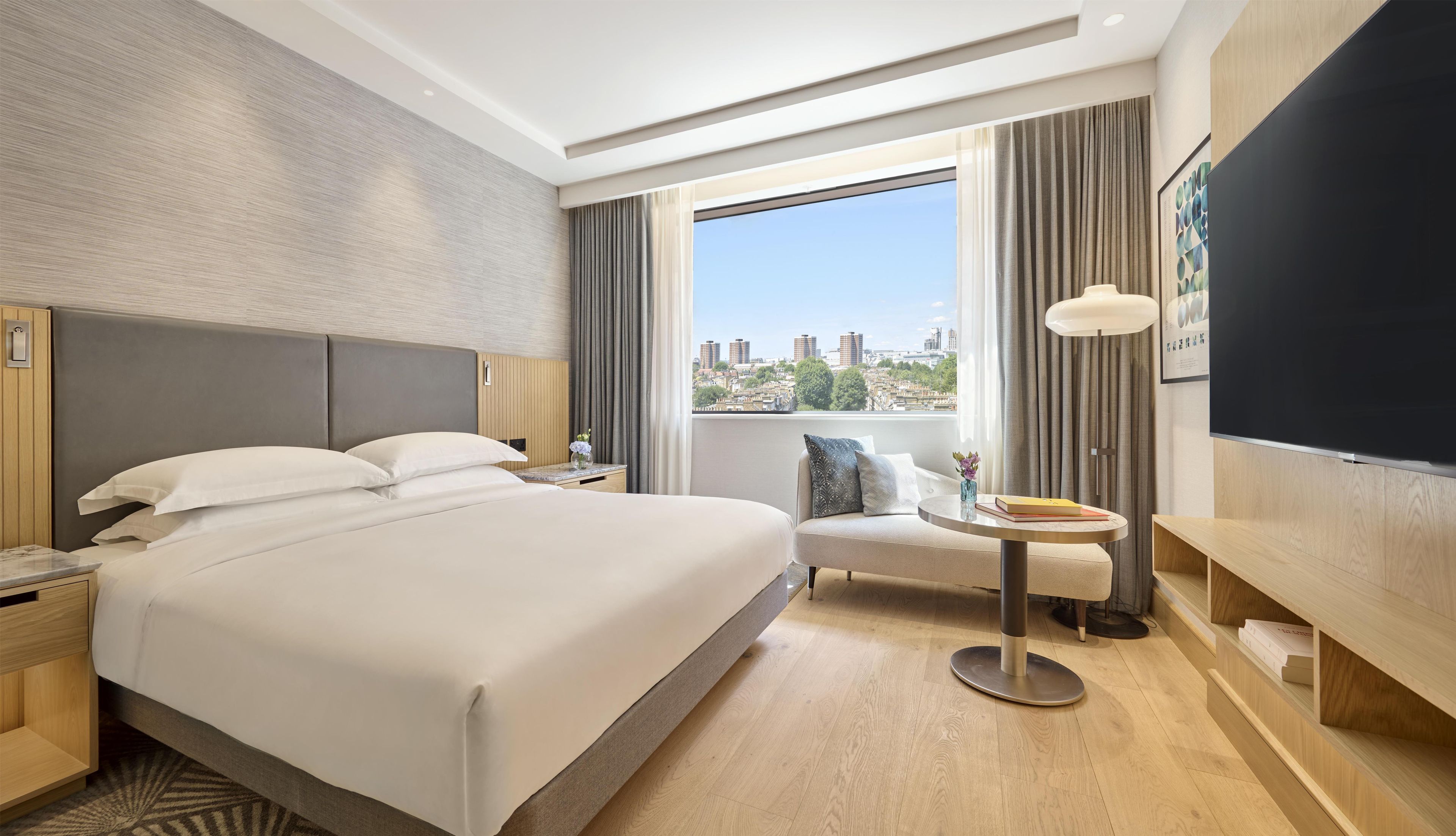 A hotel room with a large bed, a small round table, a sofa, a TV, and a window with a city view.