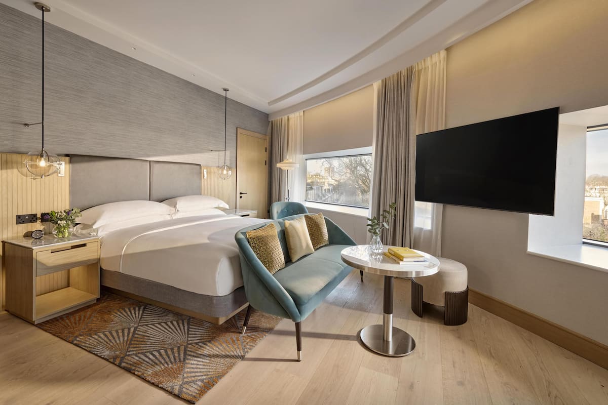A modern hotel room with a bed, bedside table, lamp, TV, and a view of the city.