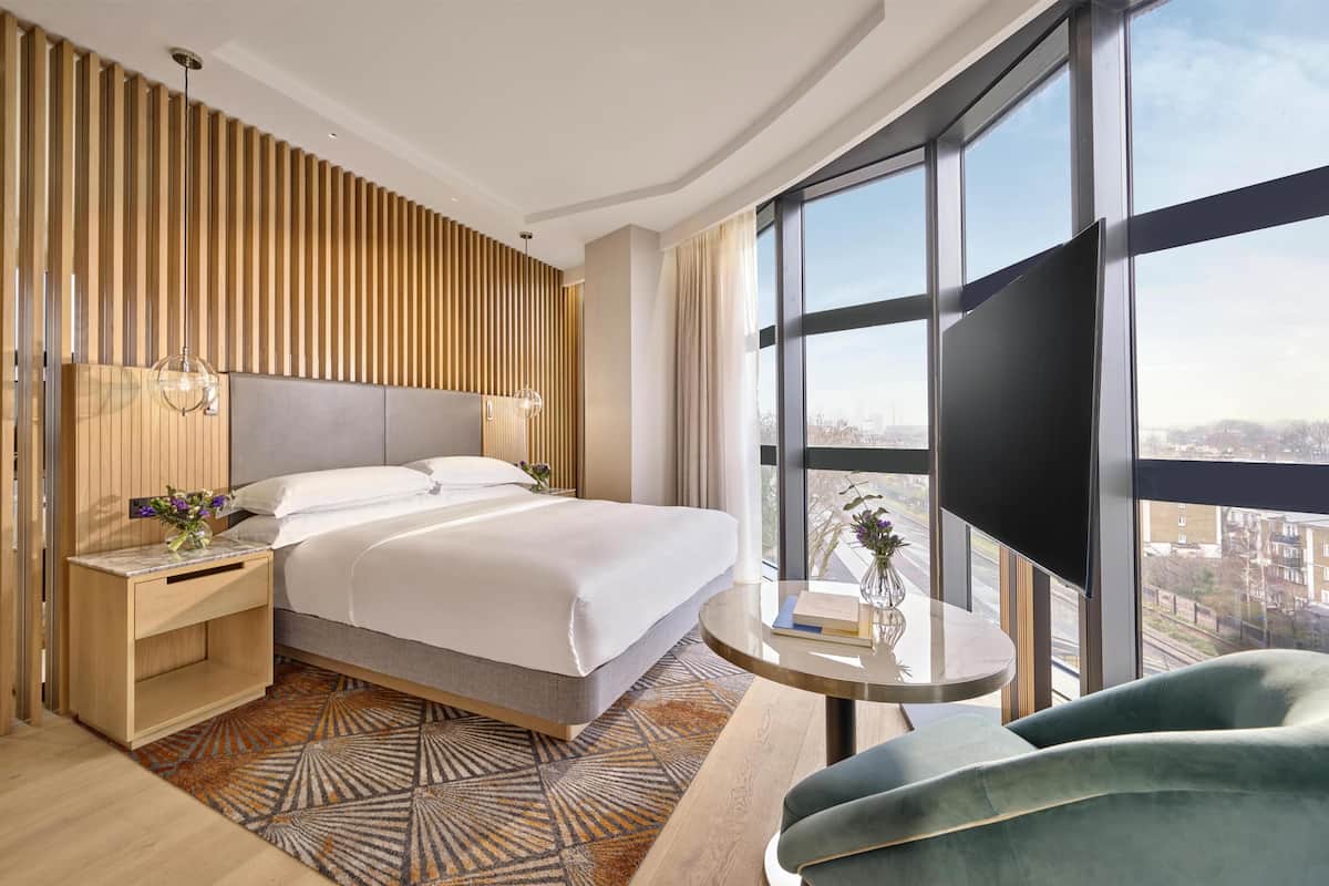A modern hotel room with a large bed, a bedside table with a vase, a round table with a chair, and a view of the city through large windows.