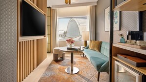 A modern hotel room with a large flat-screen TV, a sofa, a coffee table with a vase of flowers, and a view of a building through the window. - Hyatt Regency London Olympia (London)