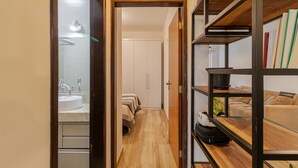 A narrow hallway with a wooden door leading to a bathroom with a sink and mirror. A bed with a white headboard is visible in the background, and a bookshelf with various items is on the right side. - Casa Pet Friendly no Condom nio Bell Park (São José dos Campos)