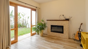 A spacious living room with a fireplace, a large window with curtains, a potted plant, and a guitar. - Casa Pet Friendly no Condom nio Bell Park (São José dos Campos)