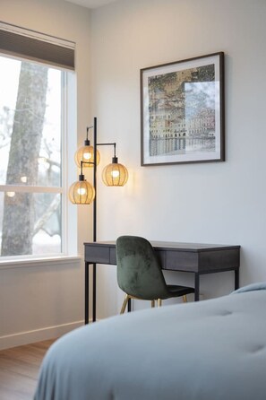 Quarto com cama, escrivaninha e cadeira. Há uma janela com persianas, um quadro na parede e um abajur.