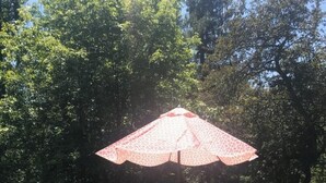 An outdoor seating area with a table and chairs under a patterned umbrella, surrounded by trees.