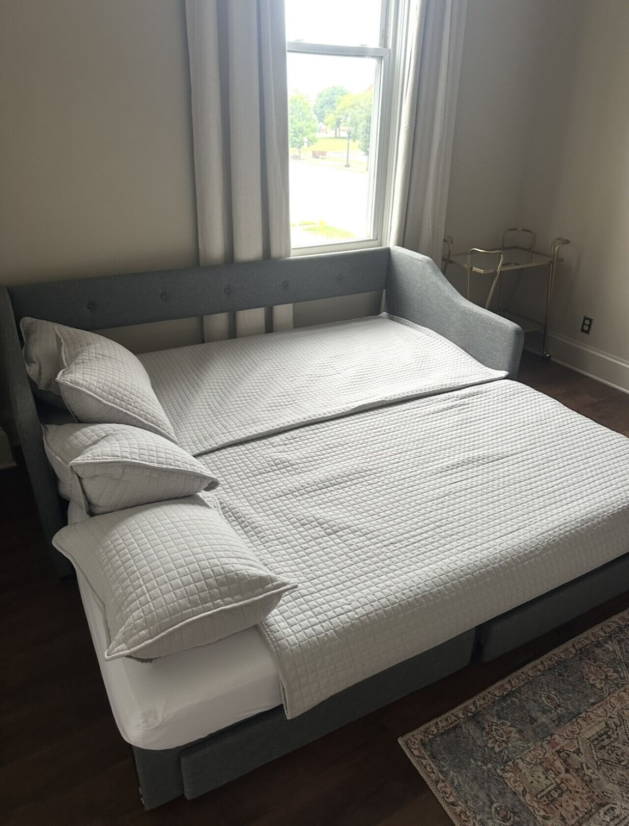 Ein Bett mit einer gesteppten Matratze und Kissen, ein Metall-Nachttisch und ein Blick ins Freie durch ein Fenster.