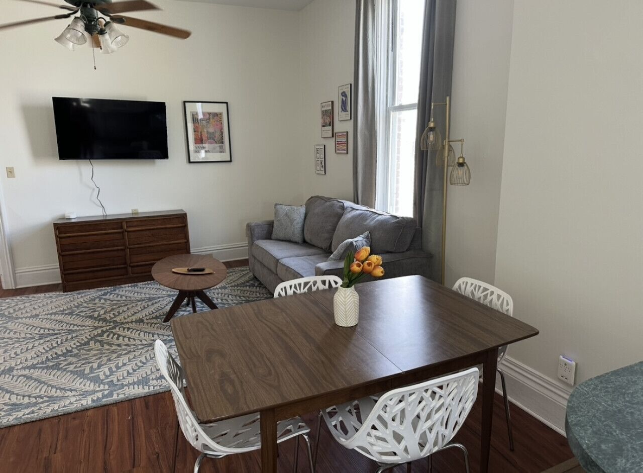 Een eetgedeelte met een houten tafel en witte stoelen, een grijze bank, een aan de muur bevestigde televisie en een plafondventilator.