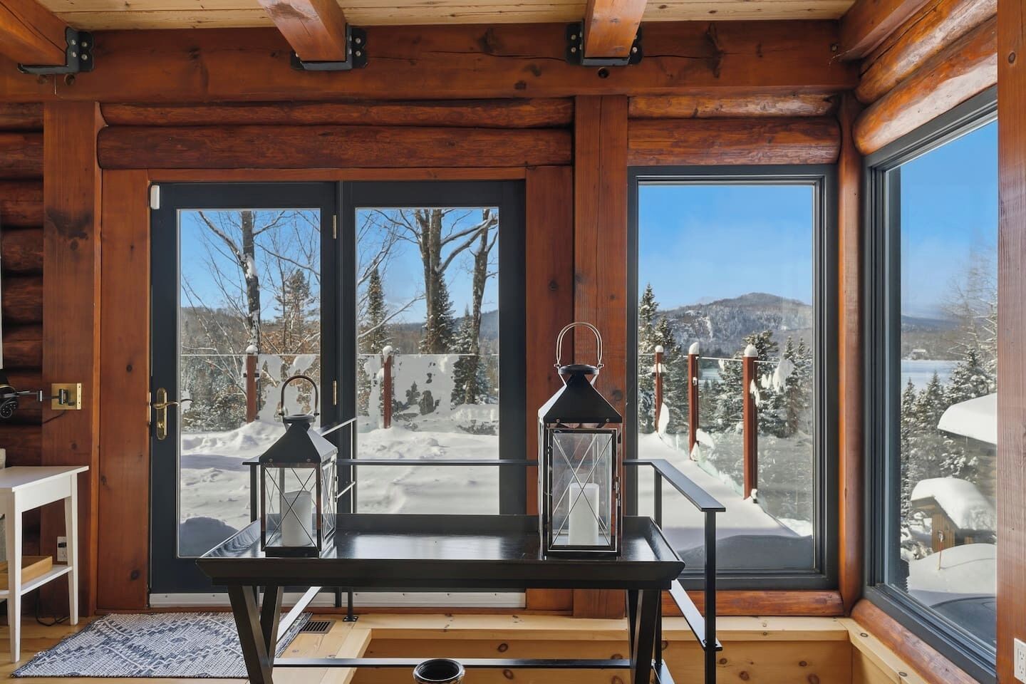 A wooden cabin interior with a table and two lanterns, a door leading to a snowy outdoor area, and a large window offering a mountain view.