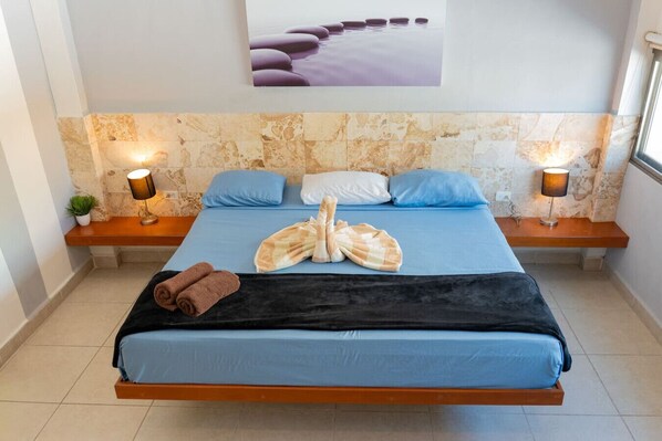 A neatly made bed with blue bedding, two bedside tables with lamps, a wall-mounted artwork, and a stone wall. - Ocean View Penthouse in the Main Center (Playa del Carmen)