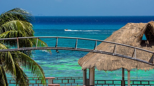 A beach view with a thatched-roof structure, palm trees, and a clear blue ocean. - Ocean View Penthouse in the Main Center (Playa del Carmen)