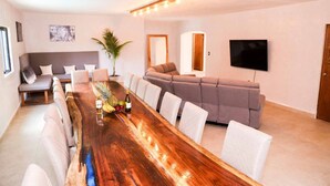 Uma sala de estar moderna com uma mesa de jantar de madeira comprida, um sofá seccional cinza e uma televisão de tela plana presa na parede.